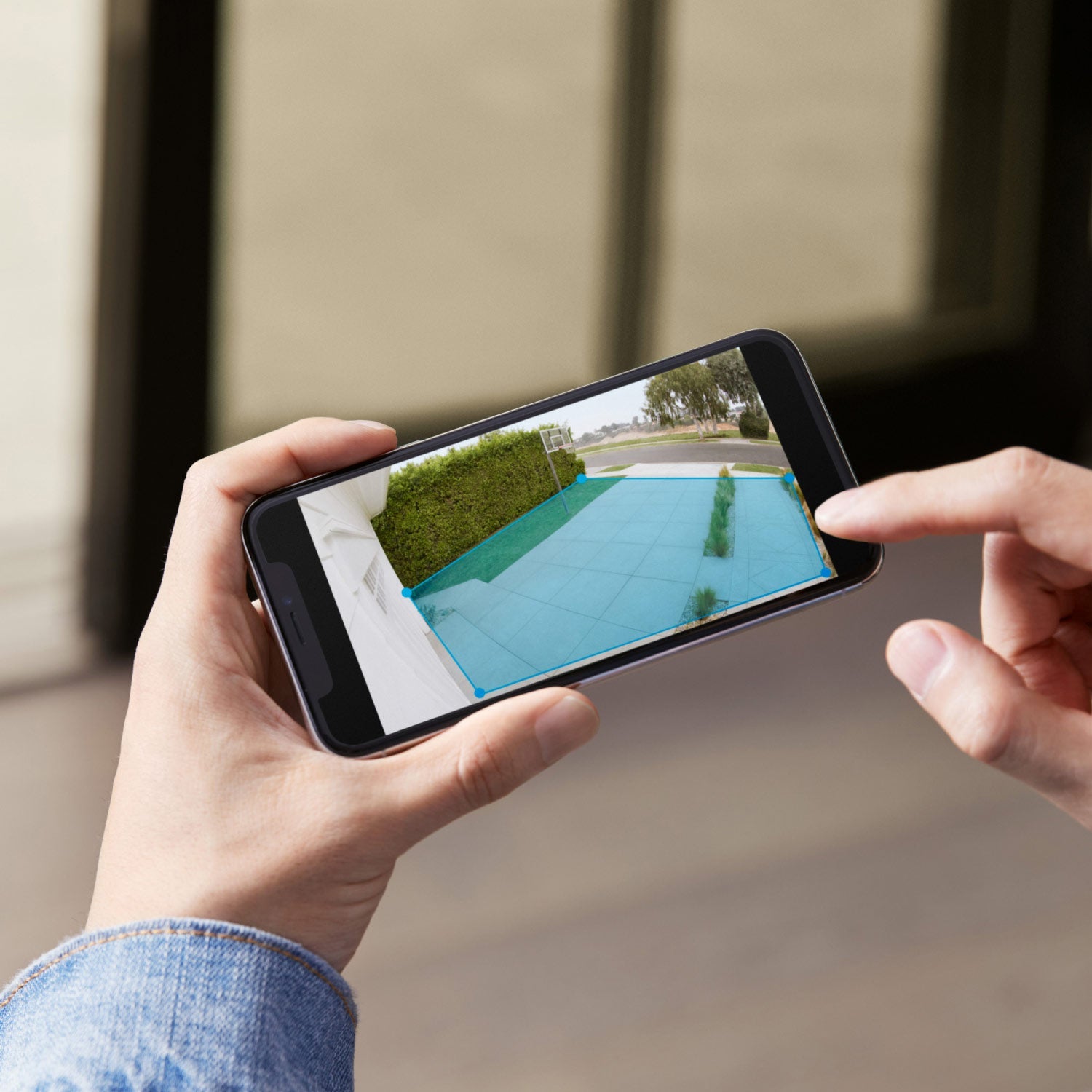 Spotlight Cam Plus - Wired:Hands holding a smartphone displaying the Ring App with customizable motion detection zone outlined in blue over a driveway and walkway in daylight.