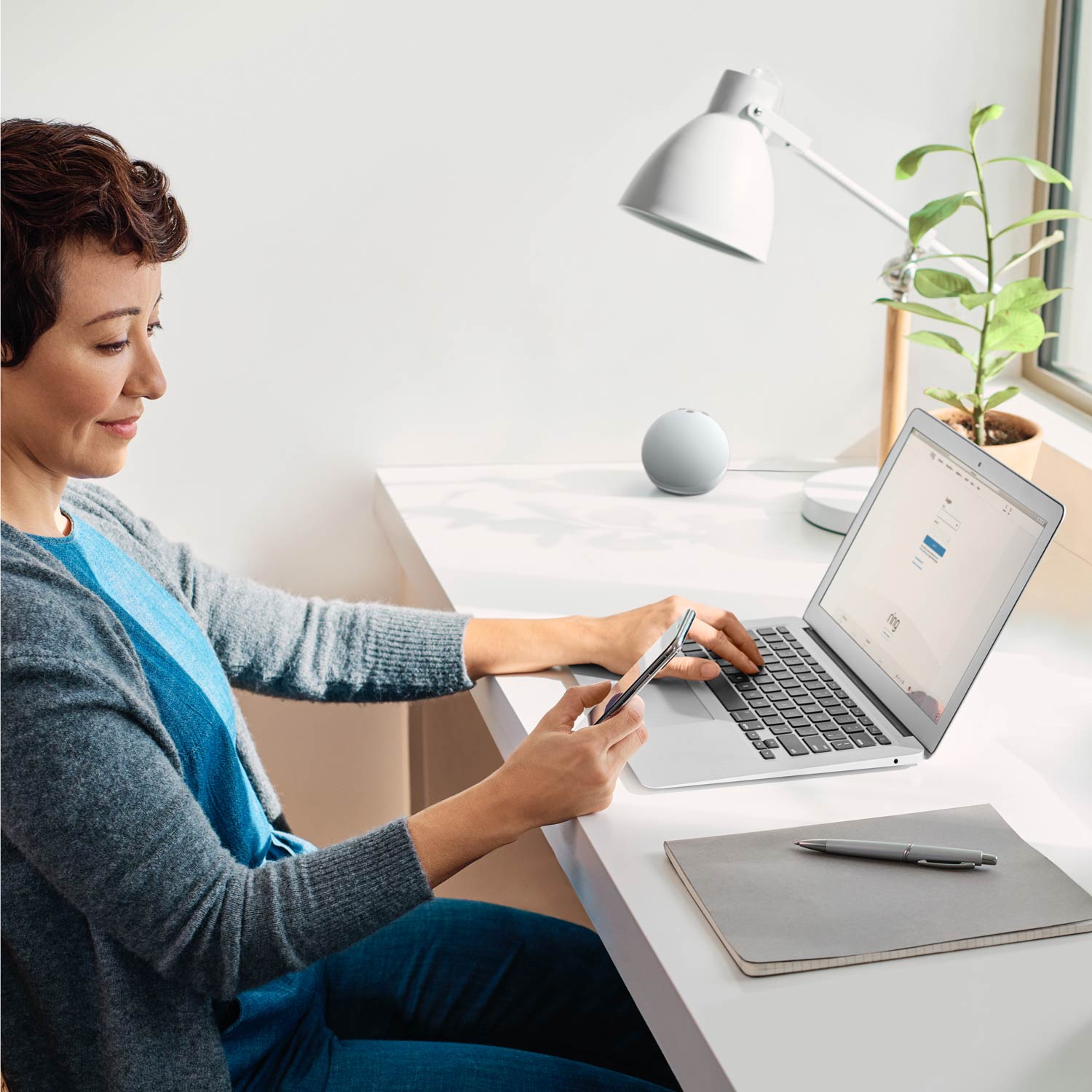 Alarm Pro, 13-Piece Kit (with built-in eero Wi-Fi 6 router and additional eero 6 Wi-Fi Router) - Woman sitting at desk using a laptop and smartphone, with smart speaker, desk lamp, and potted plant visible.