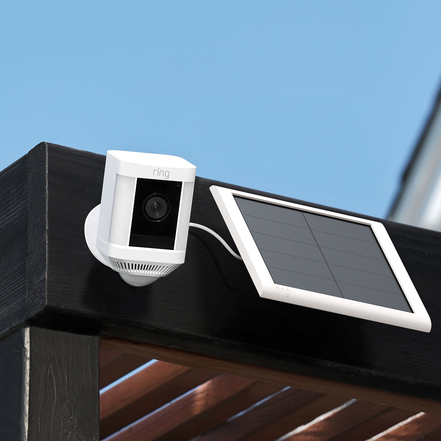Spotlight Cam Plus - Solar:White Ring Spotlight Cam Plus mounted on wooden beam, connected to white Ring Solar Panel under a clear blue sky.