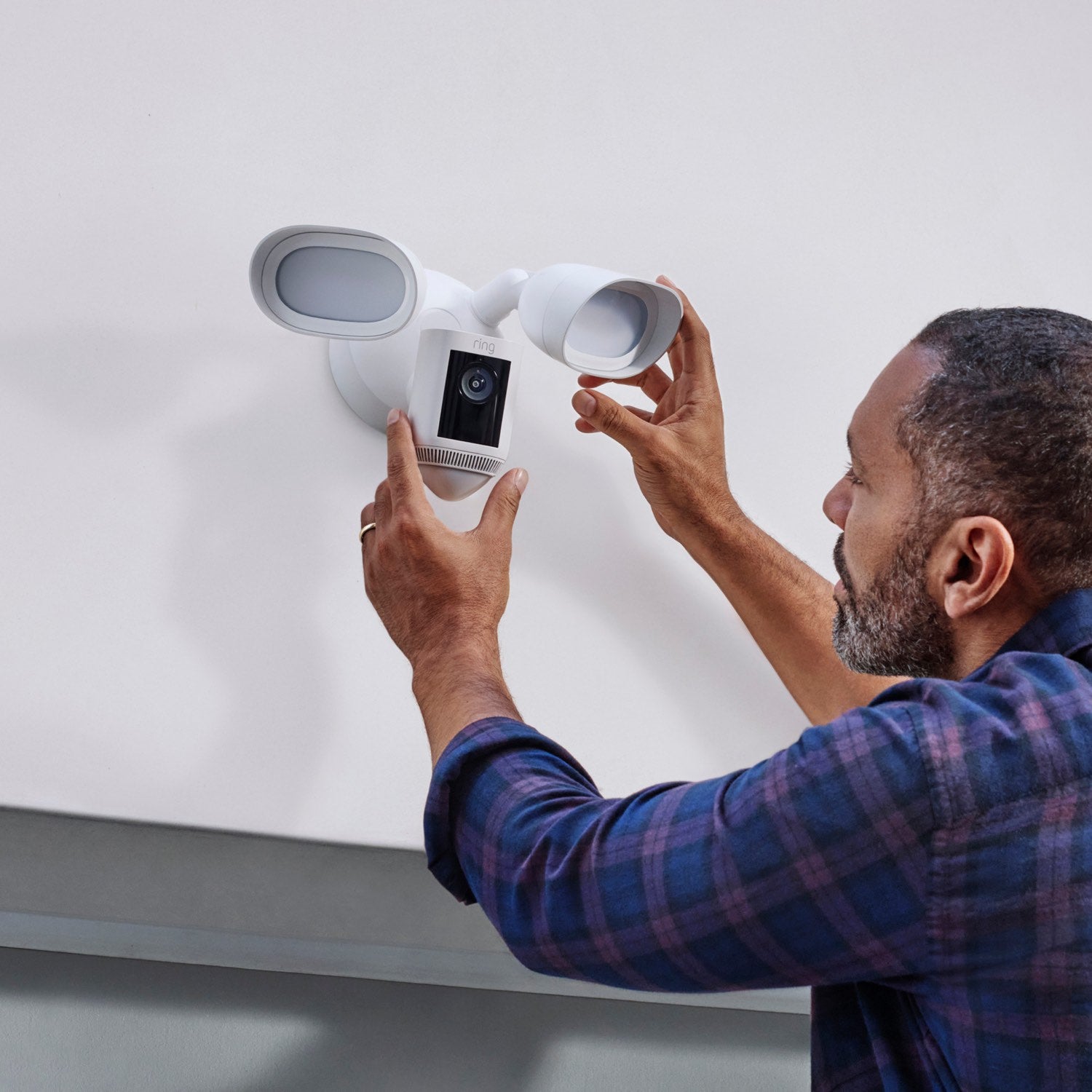 Floodlight Cam Pro (Featuring Retinal 2K) - Man wearing a purple plaid shirt adjusting a white Ring Floodlight Cam Pro mounted on an exterior wall, showing installation of camera and adjustable floodlights.
